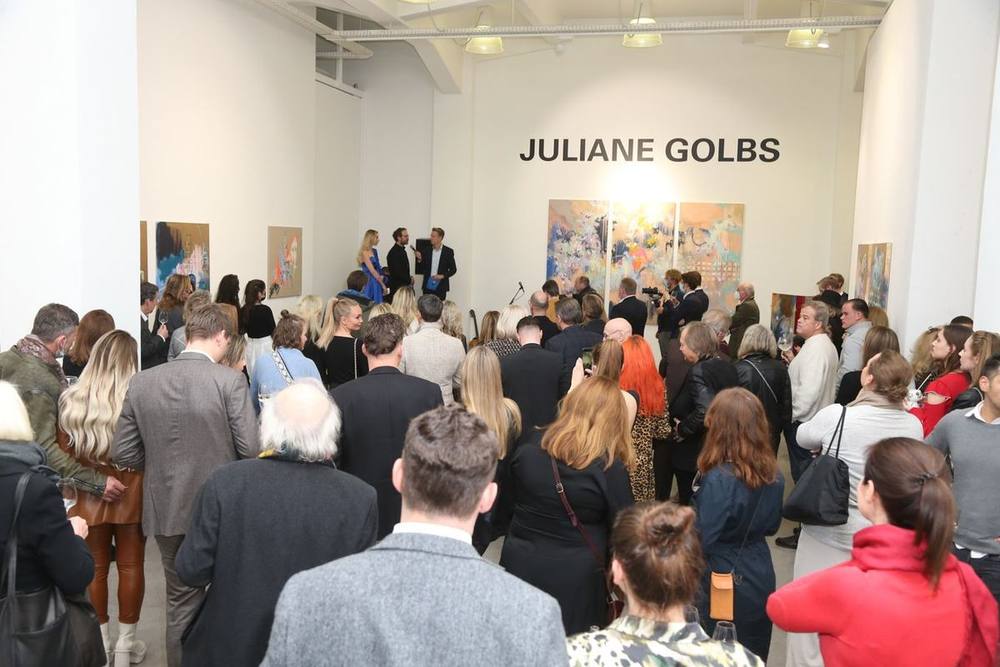 Zahlreiche Gäste bei einer Ausstellungseröffnung von Juliane Golbs, an der Stirnwand ihr Name und ein dreiteiliges Gemälde