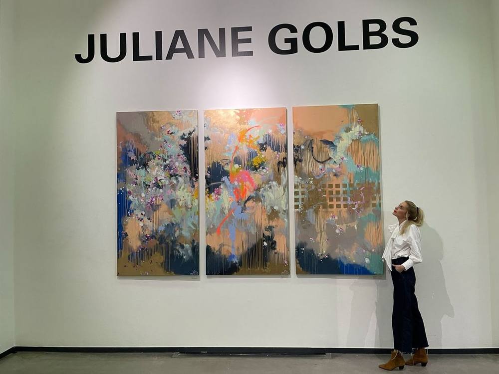 Juliane Golbs blickt auf ein dreiteiliges Gemälde in Apricot, Blau und Gold unter ihrem Namensschriftzug