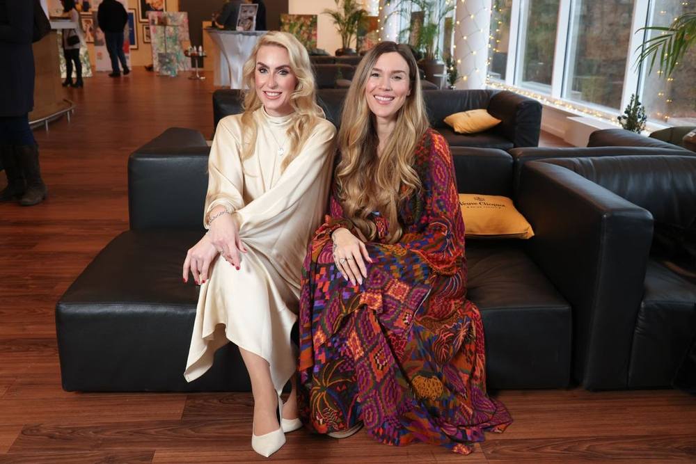 Juliane Golbs in einem cremefarbenen Seidenkleid und Joss Stone in einem farbig gemusterten Kleid sitzen nebeneinander auf einem schwarzen Ledersofa