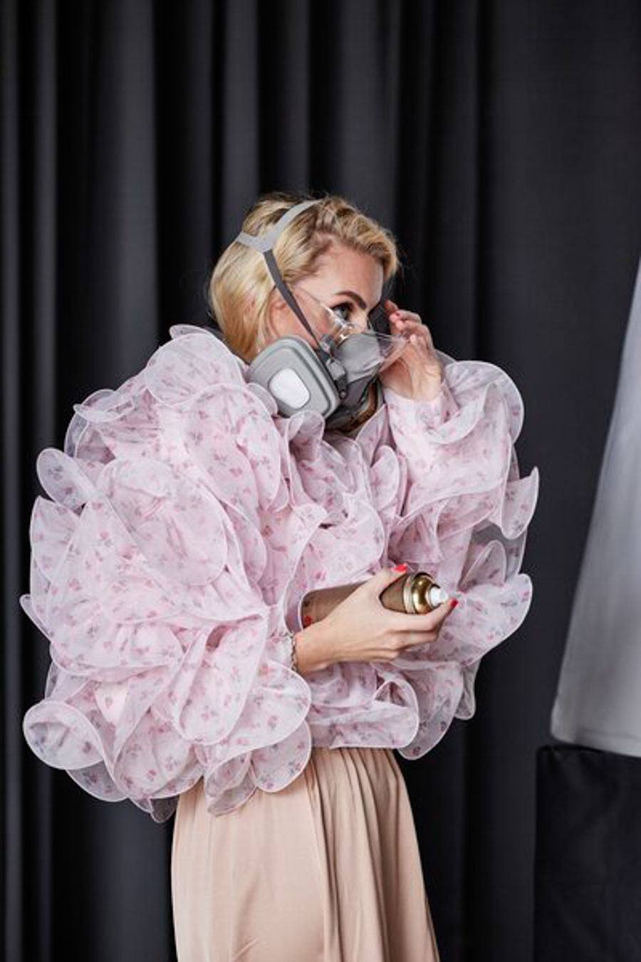 Juliane Golbs in einer rosafarbenen Rüschenjacke mit Atemschutzmaske und goldener Sprühdose